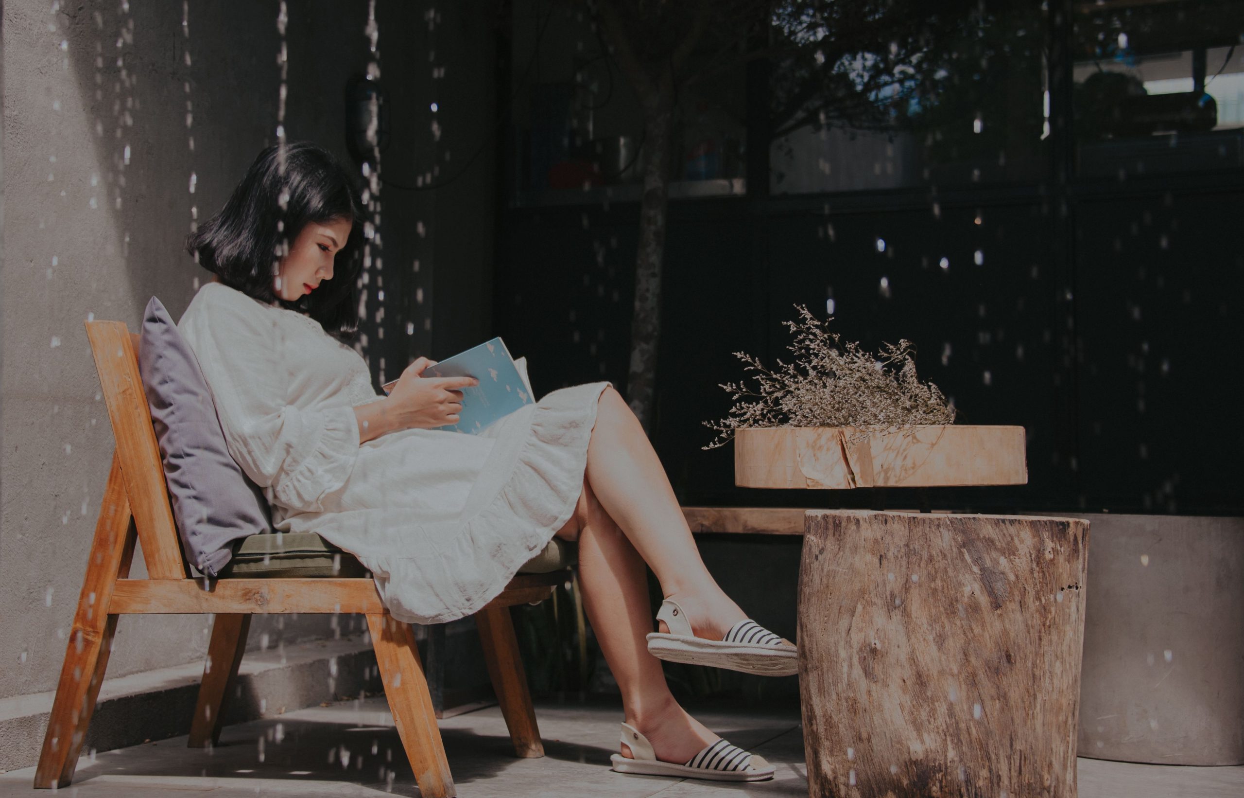 Asian woman in white dress reading in sunlight on a wooden chair outdoors.